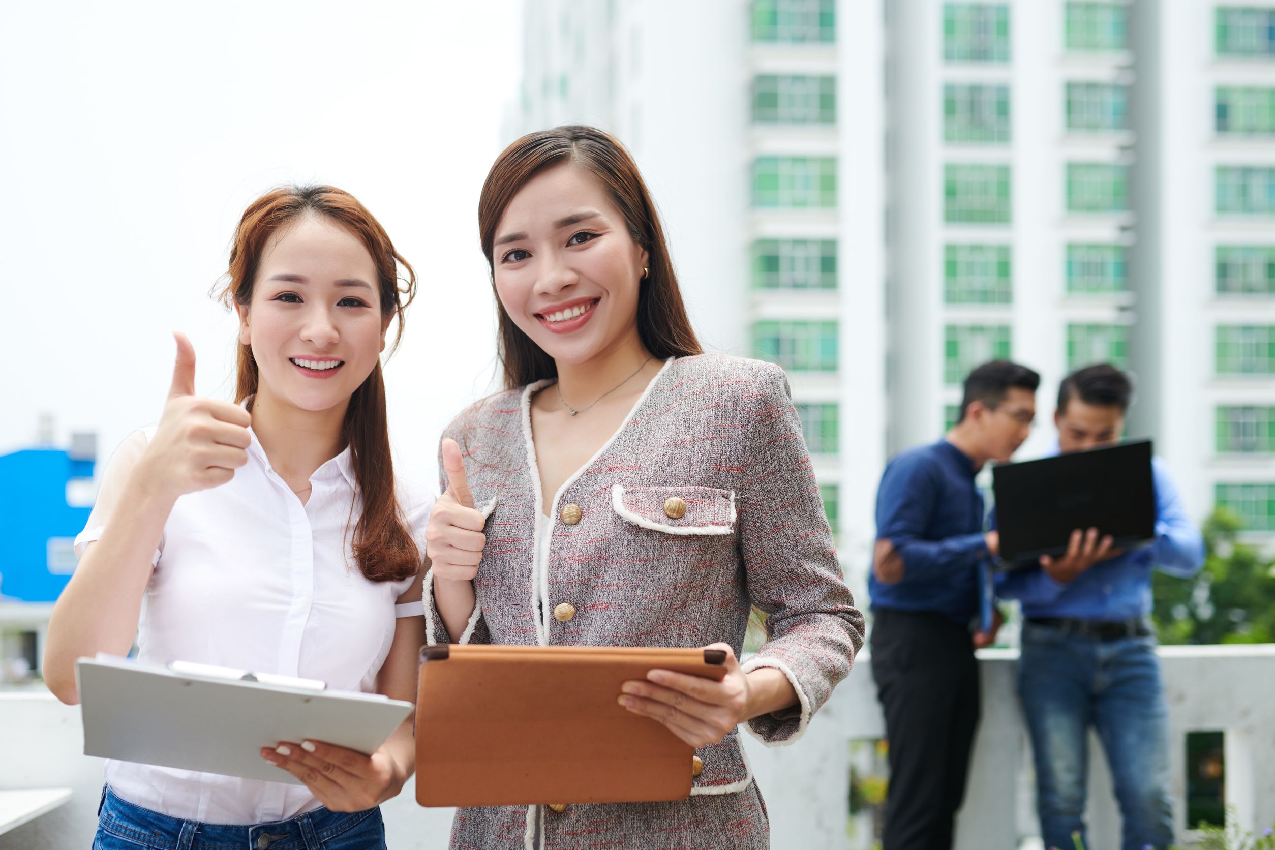 兩名年輕專業女性在戶外一同豎起拇指微笑，一人手持文件夾，另一人手拿平板電腦，背景可見兩名正在討論工作的男同事。